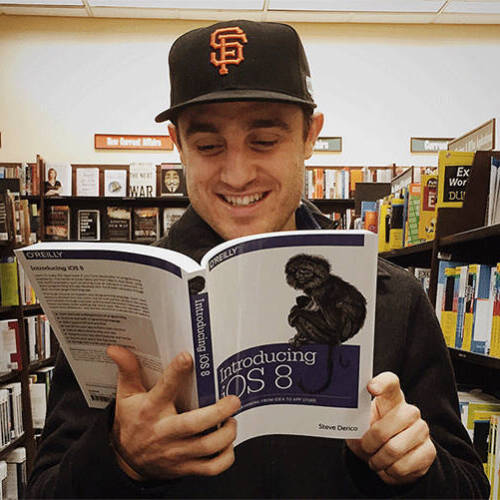 A man wearing a black San Francisco Giants baseball cap smiling while reading the book 'Introducing iOS 8' by Steve Derico in a bookstore.